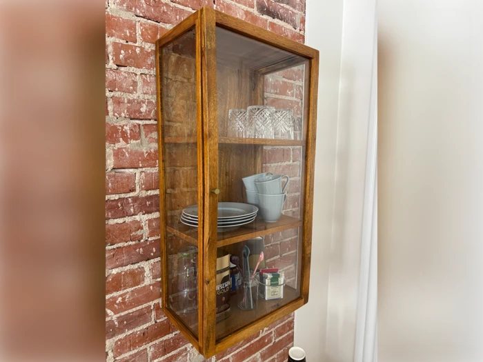 Et væghængt skab af træ og glas på en murstensvæg med glas, tallerkener, skåle, kopper og redskaber på to hylder.