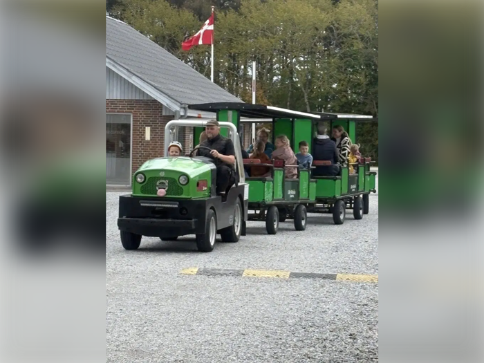 Et lille grønt tog med børn og voksne kører på grus. I baggrunden ses et dansk flag tæt på en bygning.