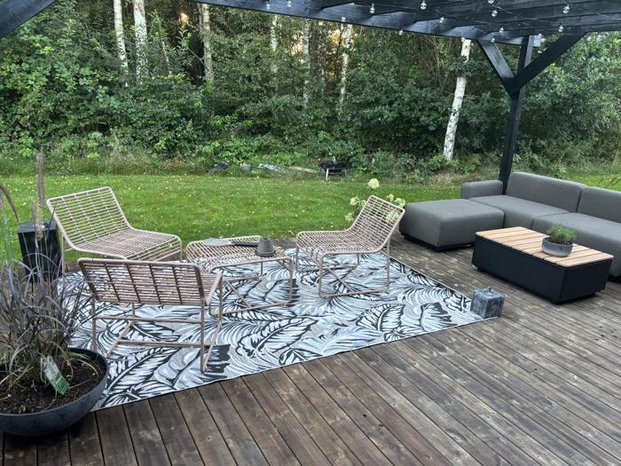 En moderne gårdhave med kurvestole, et mønstret tæppe, en grå sektionssofa, potteplanter og et trædæk på en baggrund af grønne træer og græs.