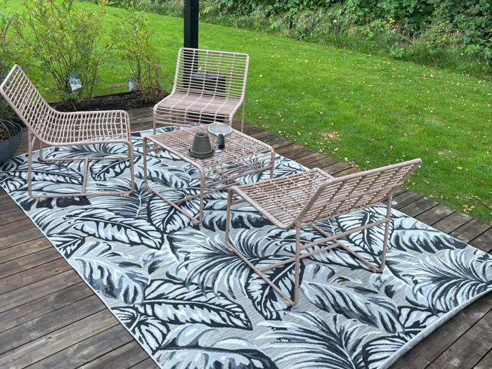 Tre kurvestole og et matchende bord står på et bladmønstret tæppe på en træterrasse med grønne planter og buske i baggrunden.