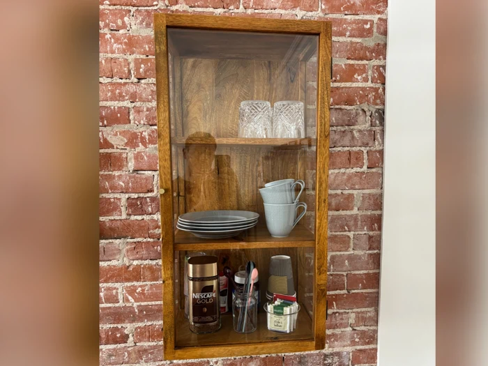 Et vægskab i træ med glasdøre op ad en murstensvæg med glas, tallerkener, krus, pulverkaffe, kopper og krydderier på tre hylder.