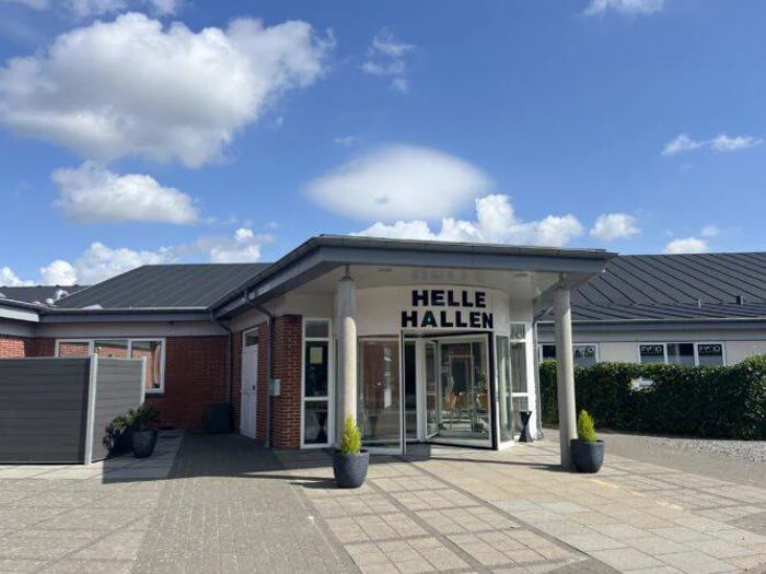 Indgang til en enetages bygning med "HELLE HALLEN"-skilt over glasdøre, to potteplanter og en blå himmel med skyer i baggrunden.