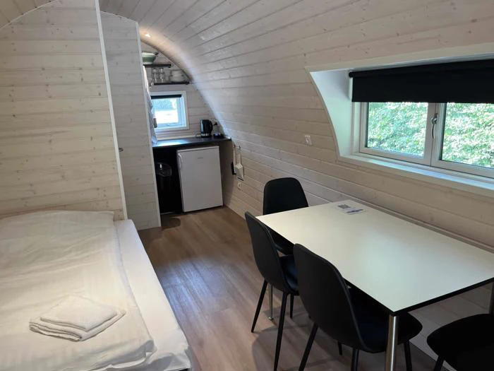 Lille hytteværelse med lyse trævægge, en seng med sammenfoldede håndklæder, et spisebord med fire sorte stole og et kompakt tekøkken nær et vindue.
