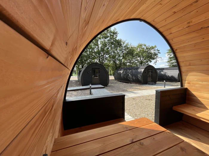 Udsigt inde fra en træhytte til tre moderne, mørkfarvede pods på en grusgrund med træer og en klar himmel i baggrunden.