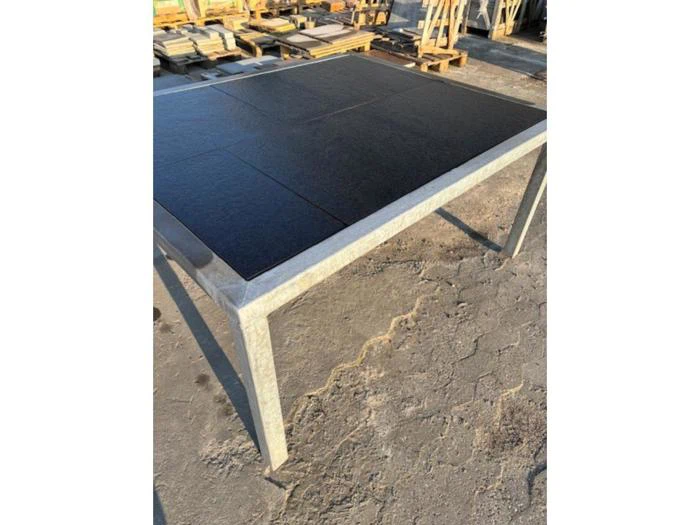 Et kvadratisk udendørsbord med metalstel og sort flisebelagt bordplade står på en asfalteret flade med stabler af materialer i baggrunden.