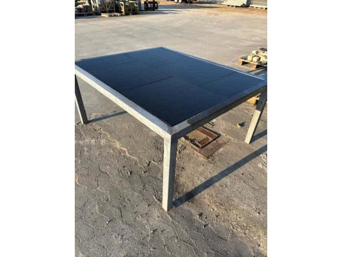 Et metalbord med en sort solpanelplade står udendørs på en asfalteret overflade i sollys.