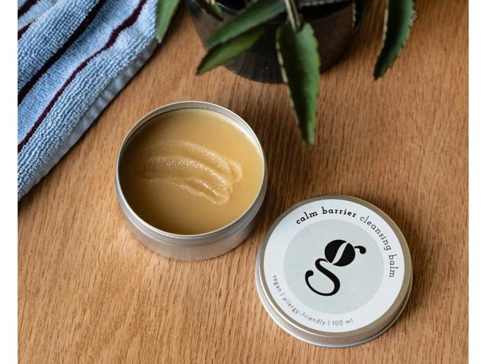 En åben dåse med Calm Barrier-rensende balsam med et cirkulært låg med mærkets logo, ved siden af en blå klud og en plante på en træoverflade.