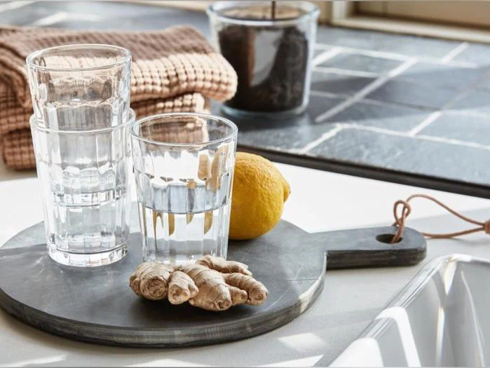 To klare glas stablet oven på hinanden, det ene glas med vand, en citron og ingefærrod på et rundt stenbræt.