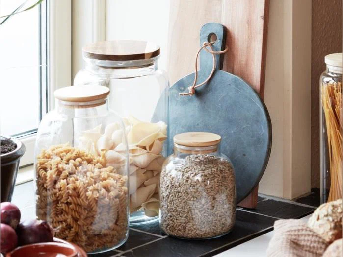 Opbevaringsglas af klart glas med trælåg, der indeholder forskellige pastatyper og korn, anbragt på en sort flisebelagt overflade ved siden af et rundt skærebræt.