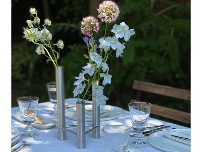 En moderne metalvase med tre rør rummer lyseblå og lilla blomster på et bord, der er dækket med et hvidt bordtæppe og klargjort til et måltid.