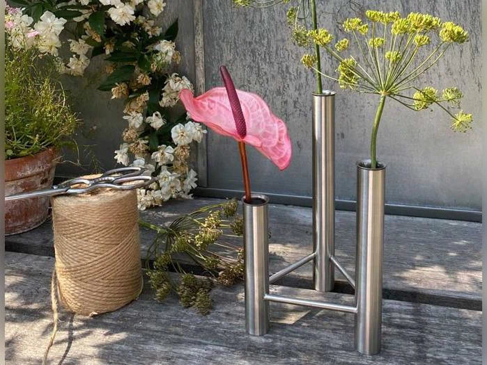 Et lyserødt anthurium og en grøn, blomstrende dildstængel arrangeret i en metalvase med tre rør ved siden af en kugle af sejlgarn.