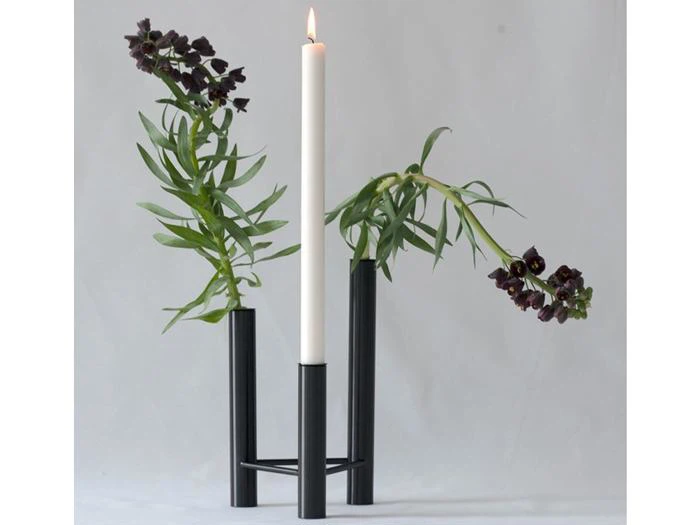 Et tændt, hvidt, konisk stearinlys i en sort metallysestage med tre rør, hvor der står mørkelilla blomster i to af rørene.