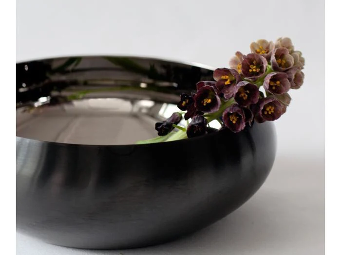 En kvist med små, mørkviolette, klokkeformede blomster, der ligger i en poleret skål af mørkt metal.