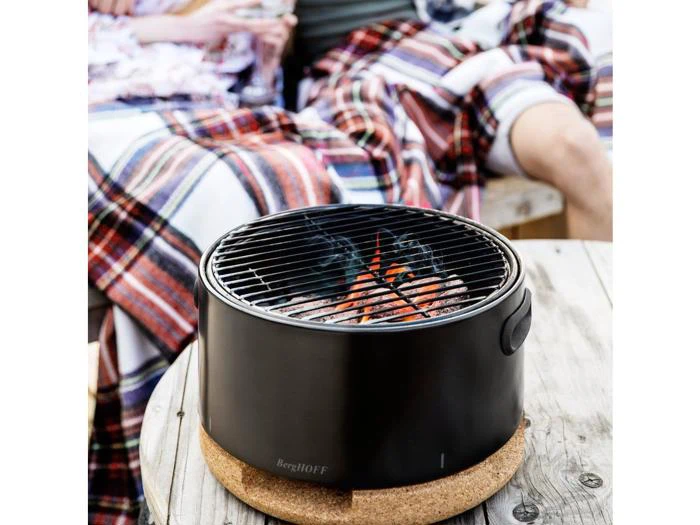 En sort, bærbar BergHOFF-grill med glødende kul står på en korkfod på et træbord, mens to personer i baggrunden er svøbt i et ternet tæppe.