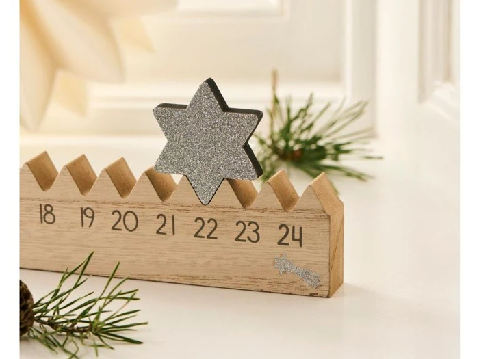 En nedtællingskalender af træ med tallene 18 til 24 har en glitrende sølvstjerne på toppen; fyrretræsgrene pryder motivet.