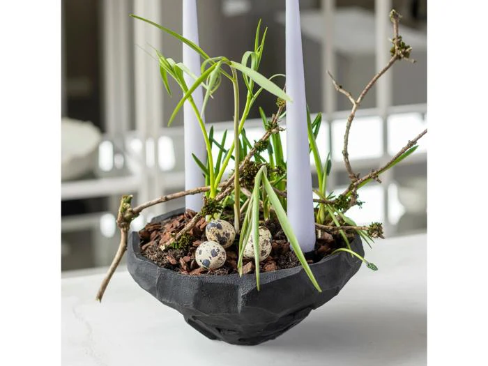 En sort skål med planter, to høje hvide stearinlys, grene, bark og tre små plettede æg, der er anbragt i jorden.