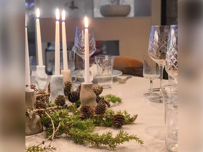 Et spisebord dækket med glas, tændte hvide stearinlys i lysestager og en borddekoration bestående af fyrrekogler og grønne grene.