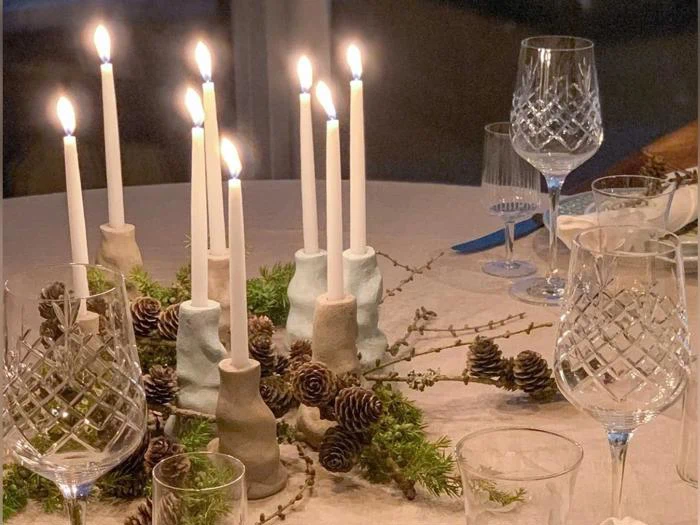 Et middagsbord med tændte stearinlys i keramiske lysestager, fyrrekogler, grønt og krystalglas, anrettet i en elegant, festlig opstilling.