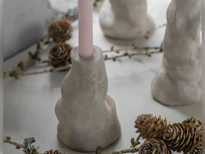 Et lyserødt stearinlys står i en struktureret, abstrakt beige lysestage, omgivet af tørrede fyrrekogler og små kviste på en lys overflade.
