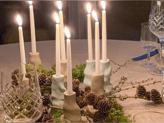 Tændte stearinlys i pastelfarvede lysestager er arrangeret på et bord, omgivet af fyrretræskogler, små kviste og grønt. Der ses også krystalglas i forgrunden og baggrunden.