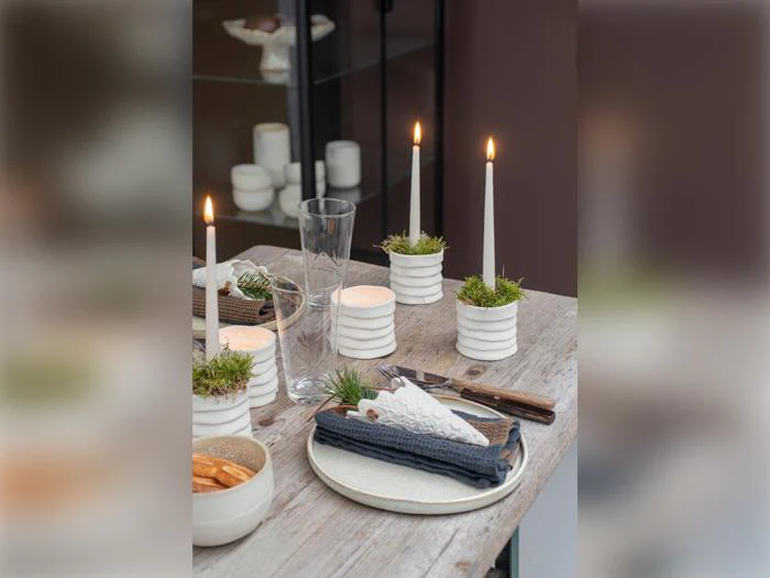 Et træbord dækket med tallerkener, bestik, en serviet, glas, småkager og fire hvide stearinlys i lysestager med mos, hvoraf nogle er tændt, hvilket skaber en hyggelig, rustik spiseatmosfære.