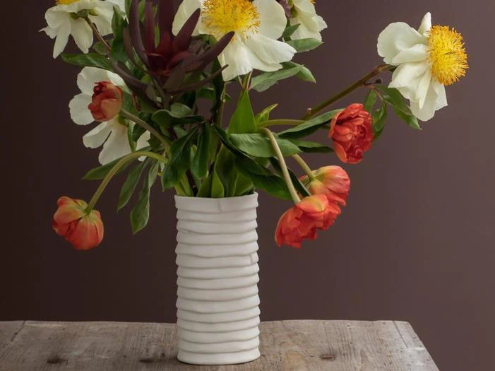 En hvid, ribbet vase rummer en buket af hvide og orange blomster med grønne blade, der står på et rustikt træbord foran en brun baggrund.