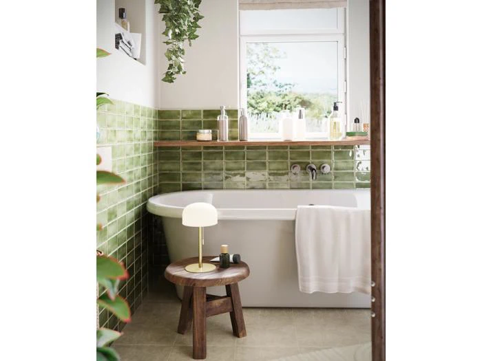 Moderne badeværelse med grønne flisewægge, et hvidt badekar, en træskammel med en lampe, toiletartikler, et håndklæde og et vindue med udsigt til grønne omgivelser udenfor.