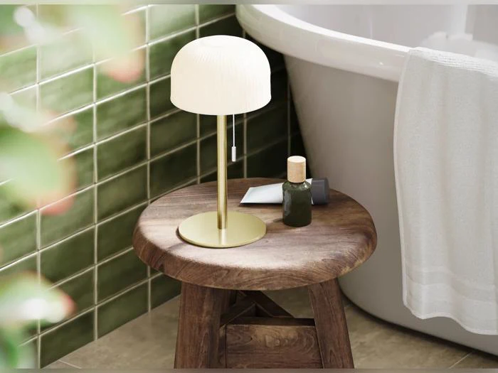 En lille lampe, en grøn flaske og toiletartikler står på en træskammel ved siden af et badekar med grønne fliser på væggene og et hvidt håndklæde hængende på kanten af badekarret.