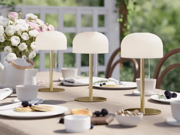 Et bord dækket til et måltid udendørs er pyntet med tre moderne hvide lamper, blomster, tallerkener, kopper, kager og bær i naturligt lys.