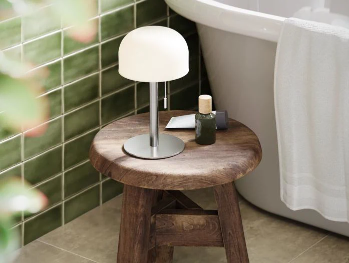 En lille rund lampe, en flaske og et håndklæde ligger på en træskammel ved siden af et badekar med grønne flisebelagte vægge i baggrunden.