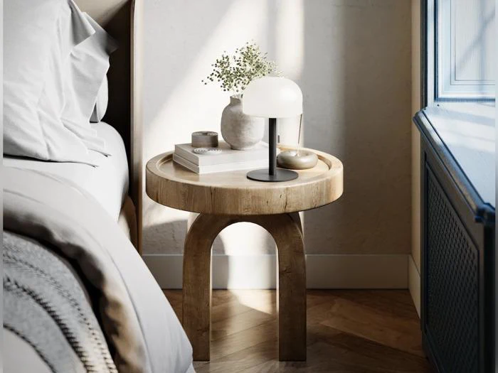 Et rundt natbord af træ med en lampe, bøger, en vase med blomster og en lille skål ved siden af en pænt redt seng i et lyst, moderne værelse.