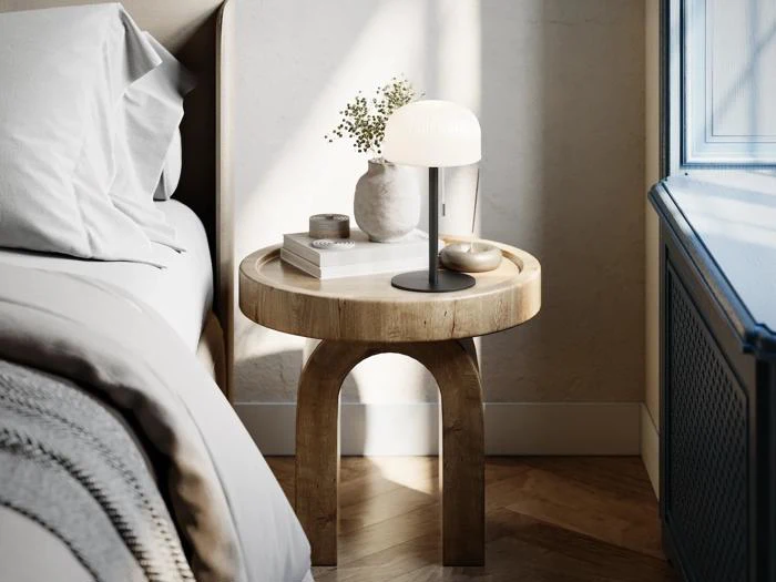 På et rundt natbord af træ står der en lampe, en lille vase med blomster, en bog og en skål ved siden af en seng med gråt sengetøj, tæt på et solbeskinnet vindue.