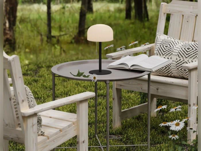 To træstole med hynder står ved et lille rundt bord, hvorpå der står en lampe, et blad, en bog og en tusindfryd, udendørs på en græsplæne med træer i baggrunden.
