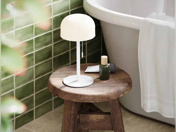 En lille hvid lampe står på en træskammel ved siden af et badekar; på væggen ses grønne fliser, og der ses også en flaske og et håndklæde.