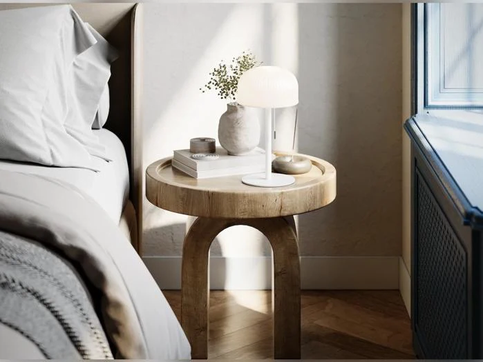 Et natbord af træ med en lampe, en vase med grønne planter, bøger og et stearinlys ved siden af en seng i et solfyldt værelse.