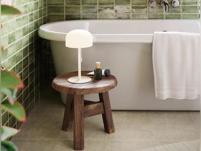 En træskammel med en lampe og toiletartikler står ved siden af et hvidt badekar med et håndklæde, foran grønne flisebelagte vægge og et beige gulv.