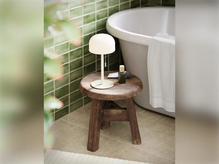 På en træskammel ved siden af badekarret står en hvid lampe og toiletartikler; i baggrunden ses grønne flisewægge og et håndklæde.