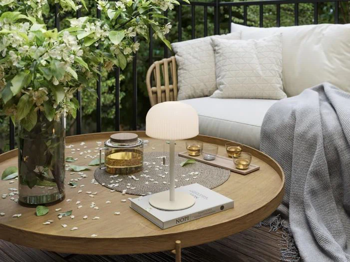 En hyggelig udendørs terrasse med et rundt træbord, en lille lampe, stearinlys, bøger, blomster og en hvid sofa med hynder og et gråt tæppe.