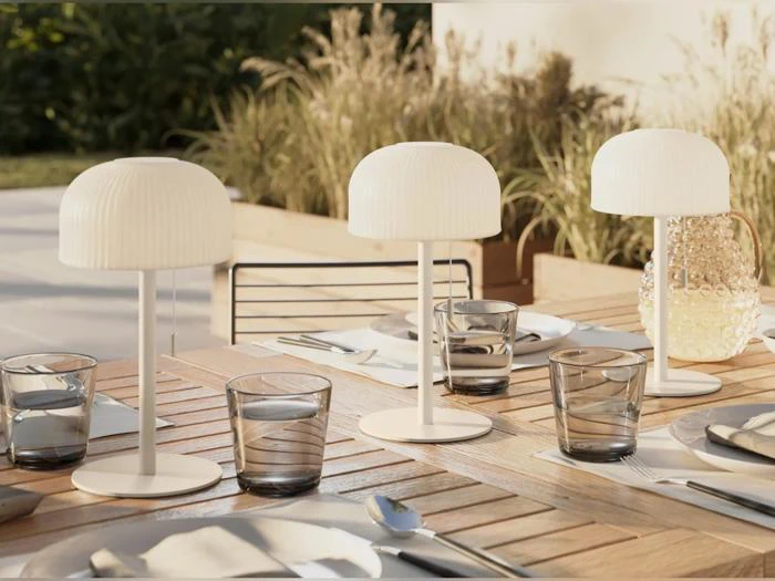 Et udendørs bord af træ er dækket med glas, tallerkener, servietter og bestik. Tre moderne hvide lamper og en kande af struktureret glas står blandt kuverterne. I baggrunden står der planter.