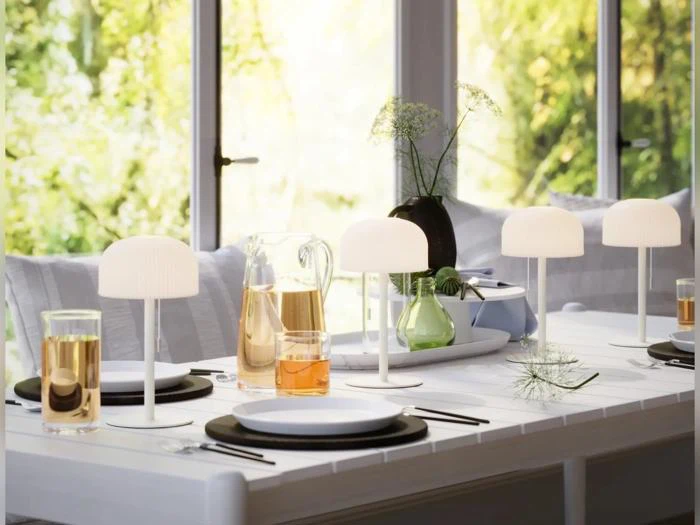Et lyst spisebord er dækket med tallerkener, glas med iste og moderne hvide lamper, med grønne planter og store vinduer i baggrunden.