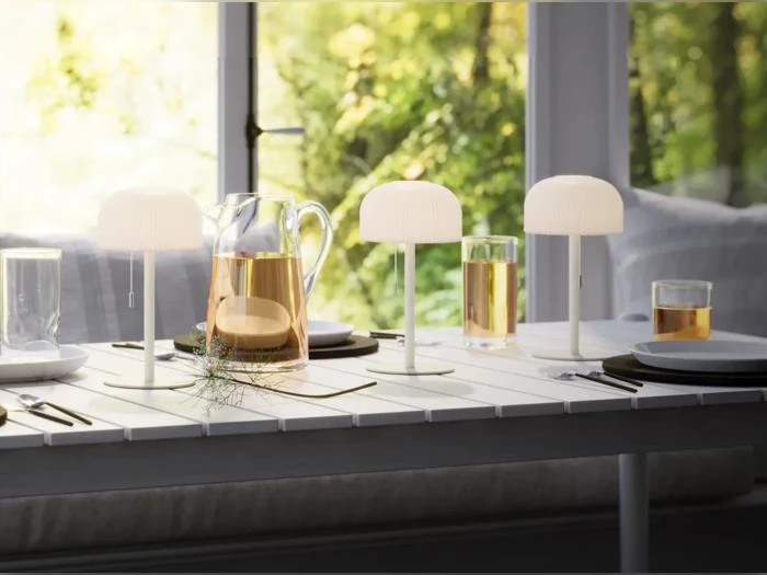 Et hvidt bord dækket med tallerkener, teglas, en glaskande og tre små hvide lamper. I baggrunden ses et vindue med grønne planter udenfor.