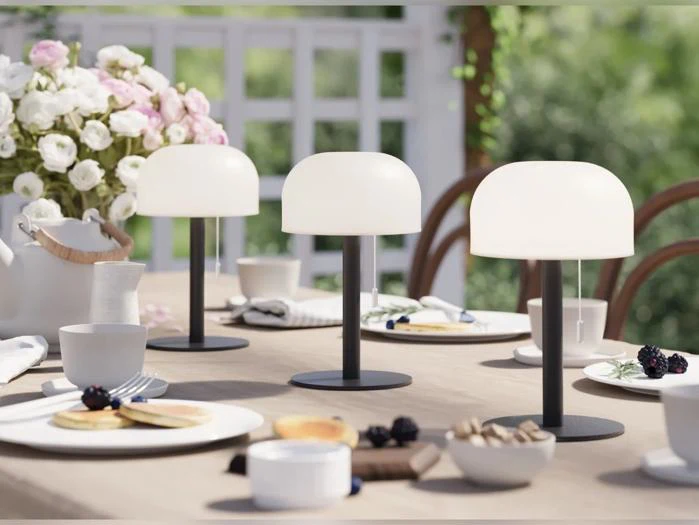 Et bord, der er dækket til et måltid udendørs, er pyntet med tre moderne hvide lamper, tallerkener med mad, kopper og en vase med lyserøde og hvide blomster.