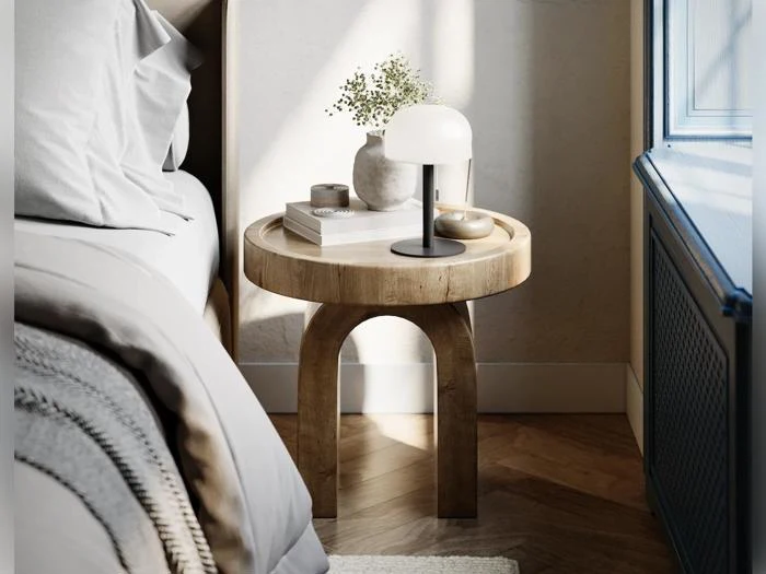 På et rundt natbord af træ står der en hvid lampe, en lille vase med grønne planter, en stak bøger og en dekorativ genstand ved siden af en seng med sengetøj i neutrale farver tæt på et vindue.