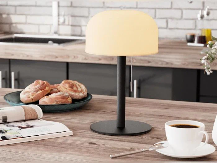 En tændt bordlampe, en kop kaffe, kager på en tallerken og et åbent magasin står på en køkkenbordplade af træ med en murstensvæg bagved.