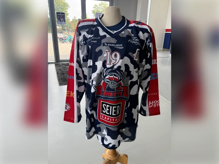 En mannequin viser en blå, hvid og grå ishockeytrøje i camouflagemønster med nummer 19, et elg-logo og flere sponsorlogoer.