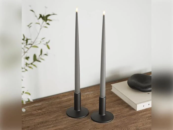 To moderne sorte, tilspidsede stearinlys med små flammer på en træoverflade, tæt ved en plante og en stak bøger med en mørk sten ovenpå.