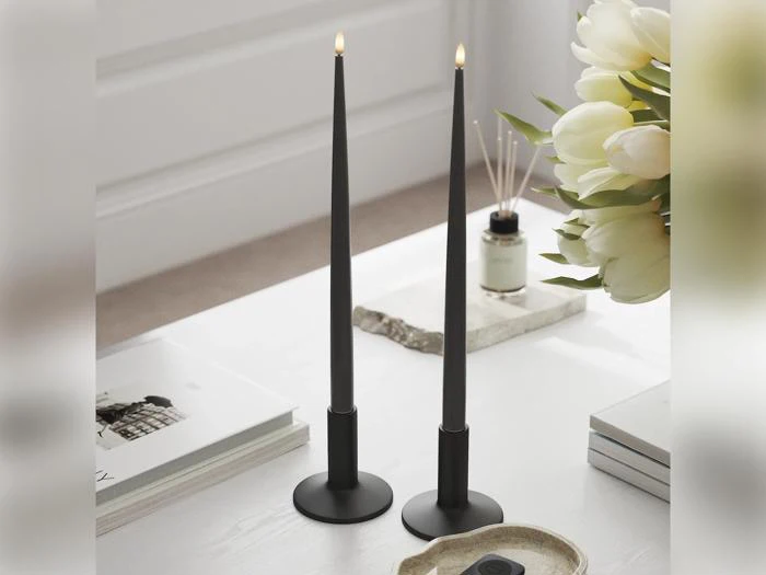 To høje, sorte, tilspidsede stearinlys med små flammer står på et hvidt bord ved siden af en vase med hvide blomster, nogle bøger og et bakke. Scenen fremstår rolig og minimalistisk.