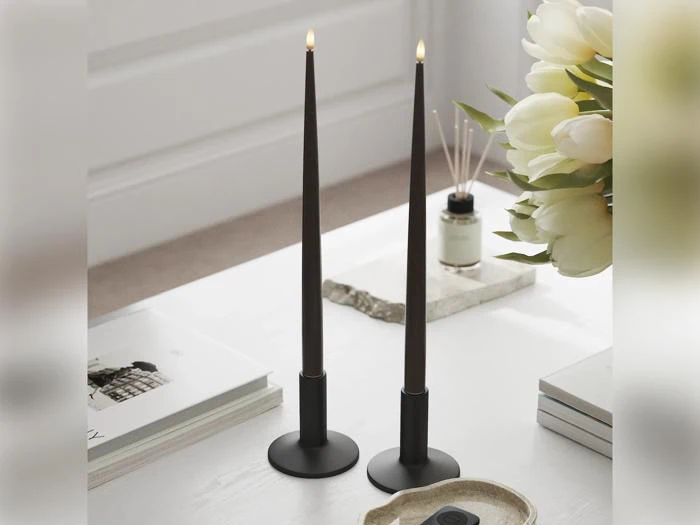 To tændte, mørke lysestager på et hvidt bord ved siden af nogle bøger, en fjernbetjening, en vase med hvide blomster og en duftdiffusor.