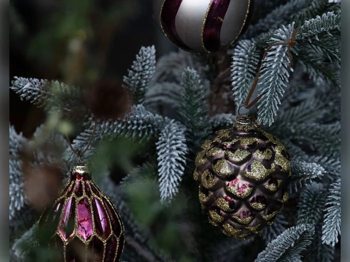 Tre dekorative pynteting, der hænger på et frostet fyrretræ: et i form af en tåre med et gittermønster, et i form af en fyrrekogle med glitter og et rundt med lodrette striber.