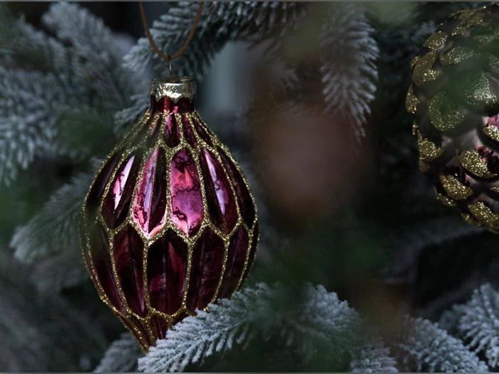 Et dekorativt, facetteret julepynt i rødbrunt og guld, der hænger på et flokket fyrretræ.